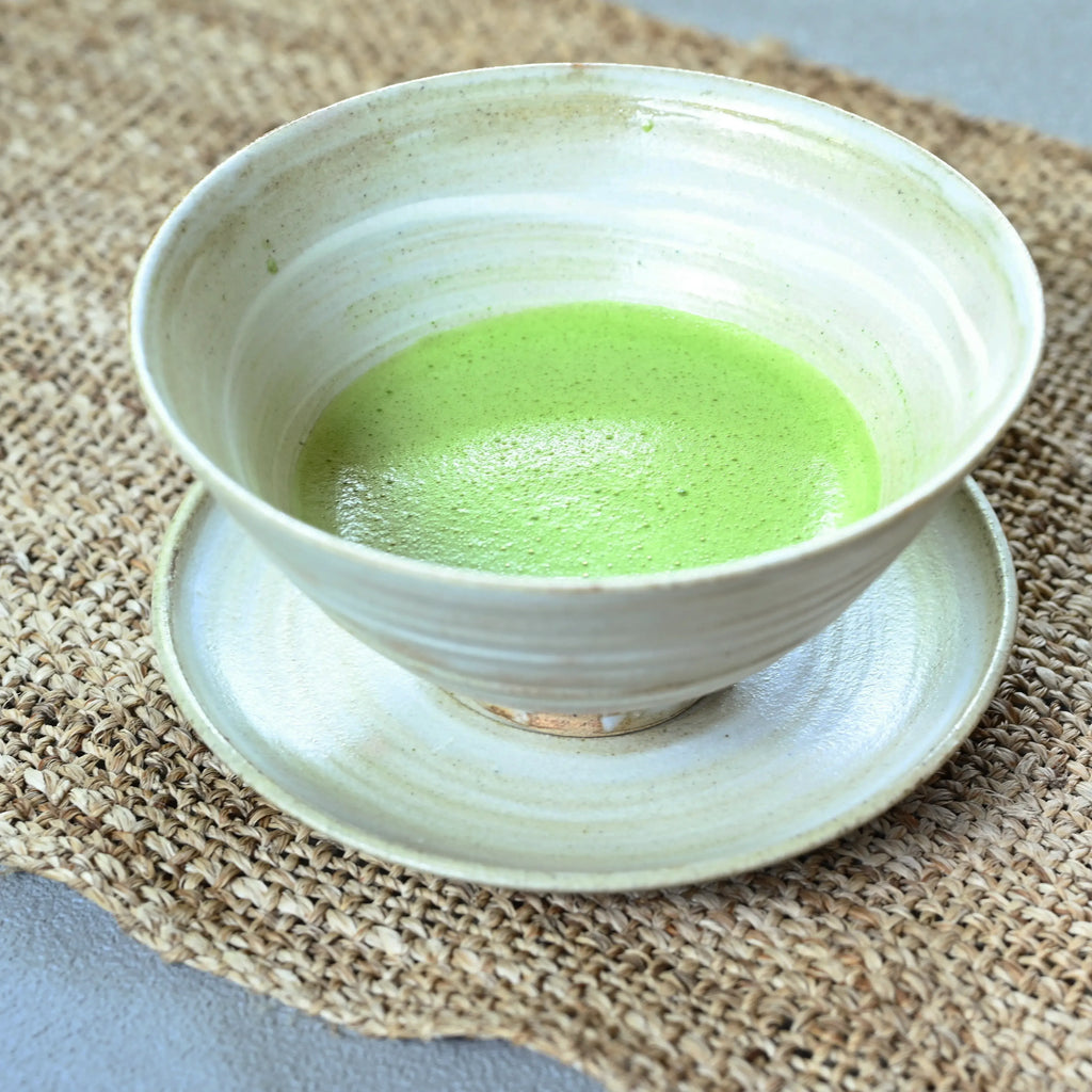A light beige ceramic bowl filled with bright green matcha tea, resting on a matching saucer over a woven burlap placemat.